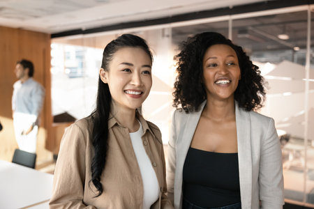 Two multiethnic businesswomen standing together in modern meeting room, smiling, exuding joy and...