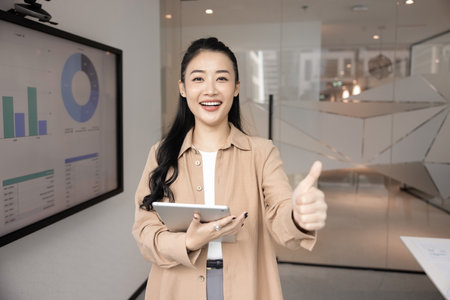Portrait of smiling Asian businesswoman analyst, project manager or sales executive holding digital tablet, look at camera, showing thumbs-up gesture. Successful project outcome, achievement, progressの写真素材