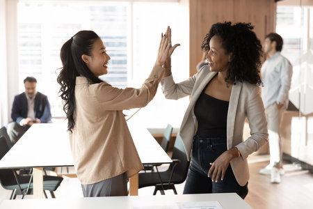 Two attractive smiling Asian and African businesswomen teammates giving high five gesture, sharing moment of joint success and mutual encouragement. Teamwork, motivation, positive working relationshipの写真素材