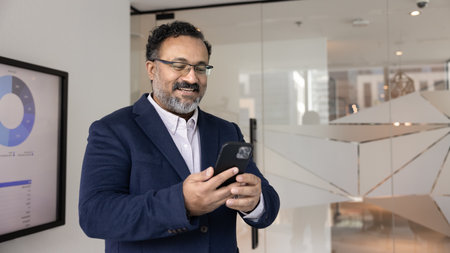 Middle-aged Indian businessman in suit using smartphone in office, reviewing documents, reading...
