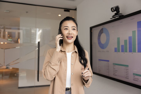 Asian professional woman engaged in phone conversation in modern office, standing next to screen displaying various charts, smiling, discussing business performance, revenue growth, or data analysisの写真素材