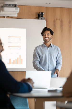 Cheerful Latino man giving presentation, stands confidently at lectern next to projection screen displays bar and pie charts with financial or budgetary data. Public speaking in modern conference roomの写真素材