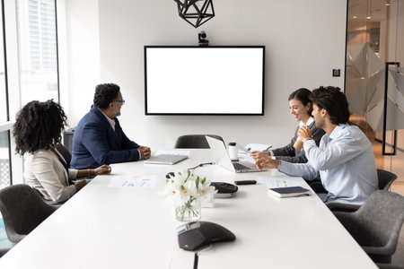 Group of four business professionals sitting around conference table in modern meeting room, looking...