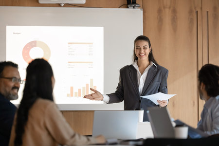 Confident businesswoman leading corporate presentation in conference room, smiling, standing beside projected whiteboard displaying financial or business-related data. Reporting, strategic planningの写真素材