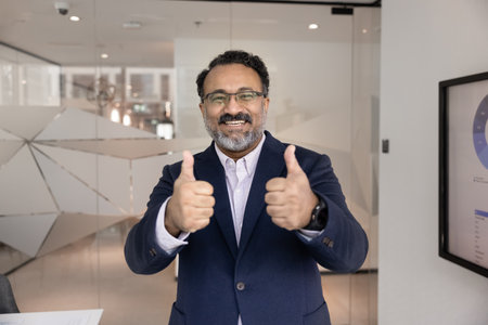 Portrait of smiling mature 45s Indian businessman in formal suit stands in office showing thumbs up celebrate great work outcome, good revenue and sales results. Business growth and financial successの写真素材