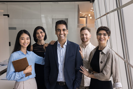 Group of friendly young employees, staff members posing for corporate photo, looking at camera with mature Indian company boss, hugging him, express bonding, showing support, unity and togethernessの写真素材