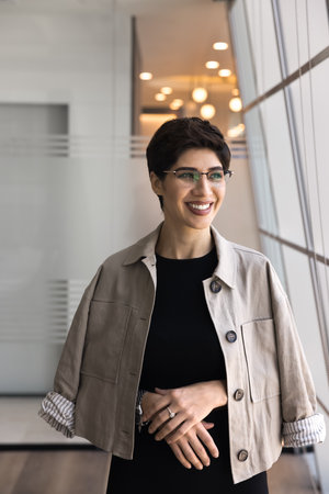 Vertical portrait of cheerful Armenian businesswoman dressed in elegant attire standing in modern...