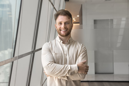Portrait of young Caucasian man dressed in casual shirt posing alone in office, standing in hallway...