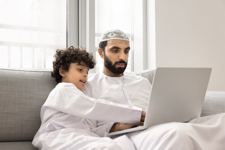 Creative and learning opportunities in modern Muslim family. Young Arab father little junior schoolboy son using notebook pc on comfy couch watch education video play game engaged in internet shoppingの写真素材