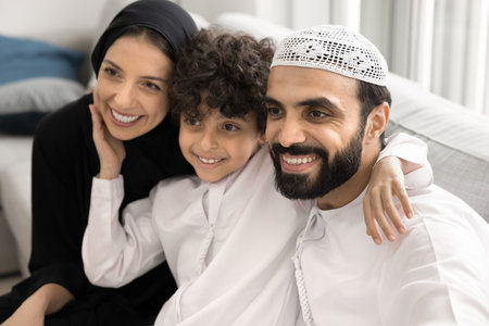 Unity in tradition and warmth in love. Family portrait happy Arabic couple with little son in faith based Islamic clothing cuddle on couch. Affectionate small boy tender touch faces of beloved dad momの写真素材