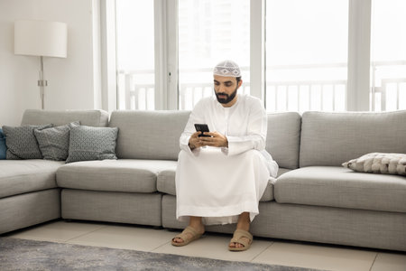 Tradition and digital tech. Young Arabian man dressed in kandora taqiyah relax on couch at modern living room using mobile phone to contact family friends scroll news text message in app. Copy spaceの写真素材