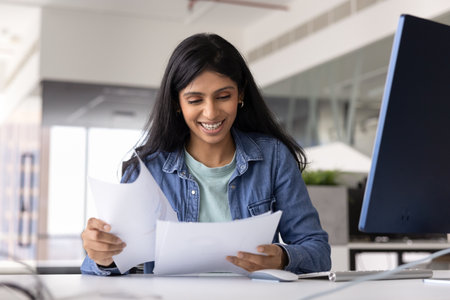 Happy young Indian marketing analyst woman reviewing sales reports at workplace with computer,...