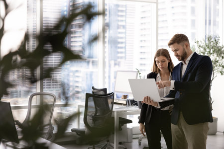 Contemporary open office workspace with two young coworkers standing together, discussing work content on laptop, using gadget for cooperation, corporate help. Candid shotの写真素材