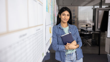 Positive young Indian company director woman in casual denim clothes posing at white board in office...