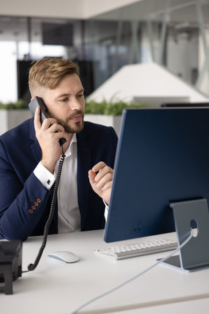 Serious busy young professional man picking up call at office workplace, talking on telephone,...