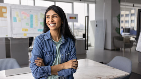 Happy successful young Indian startup owner woman in casual standing in office workspace with hands folded, looking away, smiling, laughing, thinking on successful project. Banner shotの写真素材