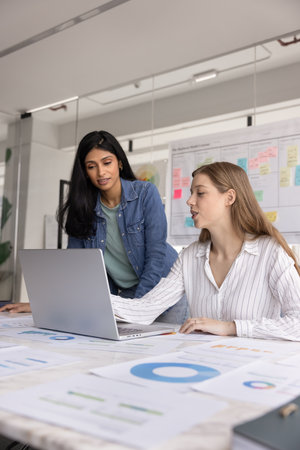 Diverse young project colleagues discussing online sales benchmarks, using laptop at table with heap of paper marketing reports, financial research results, pointing at screen, talking. Vertical shotの写真素材