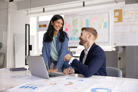 Cheerful young Indian project manager woman talking to male office coworker, smiling, discussing...