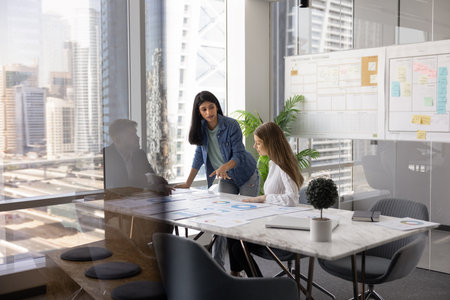 Diverse team of young startup managers and company director meeting in collaboration room at large table behind glass wall, talking, discussing paper marketing reportsの写真素材