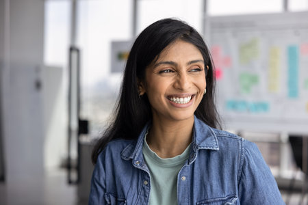Cheerful young Indian business startup leader woman posing for portrait in office, looking away with...
