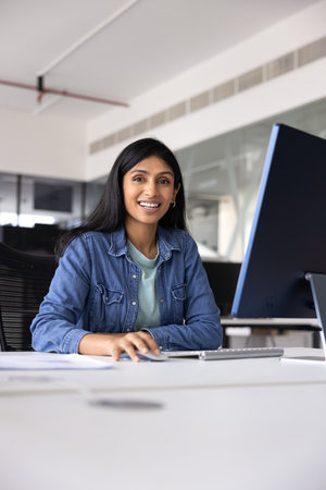 Positive young Indian product owner woman working at computer in office, posing for corporate portrait at office table with monitor, looking at camera, smiling. Vertical shotの写真素材