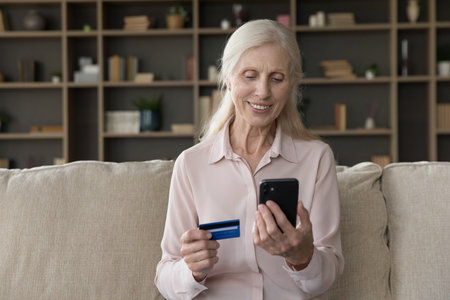 Financial operations online. Smiling confident older woman sitting on couch using credit card phone...