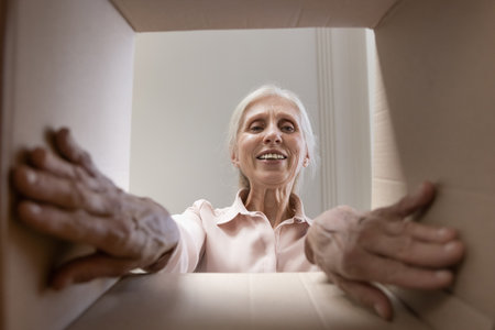 What is inside. Curious interested smiling senior woman open unpack cardboard box package with postal courier delivery enjoy online order unboxing process ready to take out surprise gift. Bottom viewの写真素材