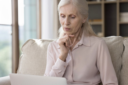 Thinking on information. Thoughtful pensive older age female retiree sit on sofa concentrated on...