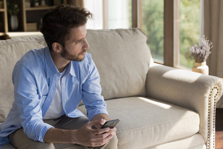 Pensive young man spend leisure time with phone at cozy home apartment sit on couch look away distracted from message text on screen create answer to chat plan online order wait for call. Copy spaceの写真素材