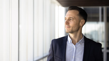 Positive young Hispanic executive man standing in office hall, looking at large window, thinking on...