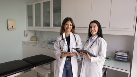Two happy successful Latin doctor women in lab coats standing together in clinic room, meeting for teamwork, professional consultation, support, looking at camera, smiling for portrait. Banner shotの写真素材