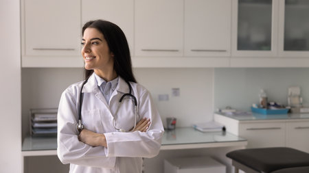Happy young Hispanic medical professional woman keeping hands folded in confidence gesture, standing...