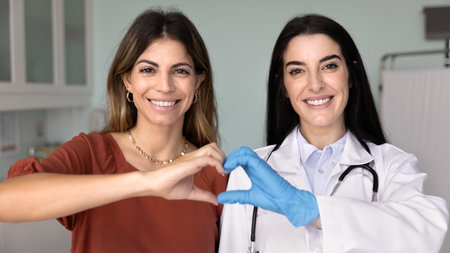 Happy cheerful young Latin doctor and patient women joining fingers, showing hand heard shape, symbol of healthcare, medical support, charity, donation, looking at camera, smiling. Banner portraitの写真素材