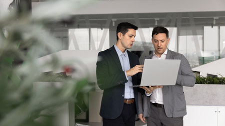 Two multiethnic men dressed in formal suits analyzing information displayed on laptop standing in...