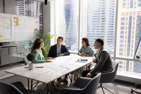 Four multinational business partners gathered at conference table to discuss joint project details or updates, strategizing together in modern office view city view. Teamwork, cooperation, negotiationの写真素材