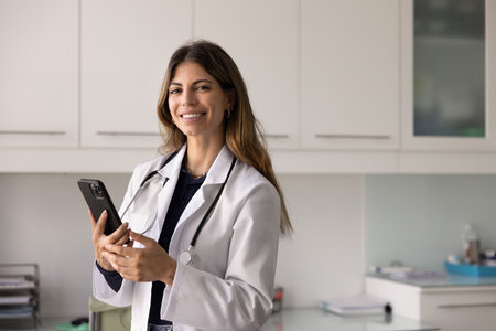 Happy successful female Hispanic doctor woman holding mobile phone, posing I hospital office for medical professional portrait, looking at camera, smiling, using gadget for online consultationの写真素材