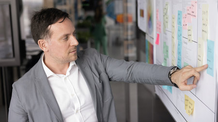 Focused professional man brainstorming, standing in front of large planning board filled with sticky...