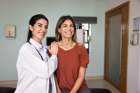 Cheerful young Latin doctor touching shoulders of female patient in supportive care gesture, looking...