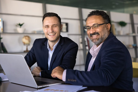 Two happy diverse younger and elder businessmen posing for professional portrait at workplace with laptop, looking at camera, smiling, symbolizing different aged teamwork, partnershipの写真素材
