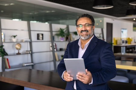 Happy mature Indian CEO man in formal jacket and eyeglasses posing in office co-working hall for...