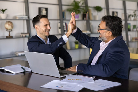 Two young and senior multiethnic businessmen making fist bump gesture at workplace, smiling,...