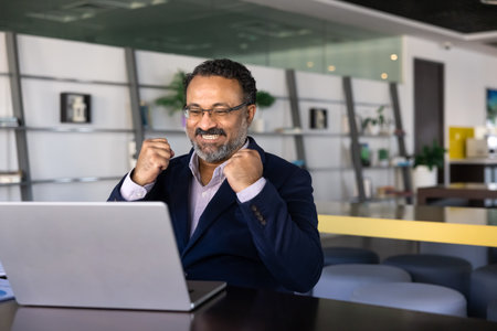 Joyful excited senior Indian online product owner making winner yes fists at computer, celebrating...
