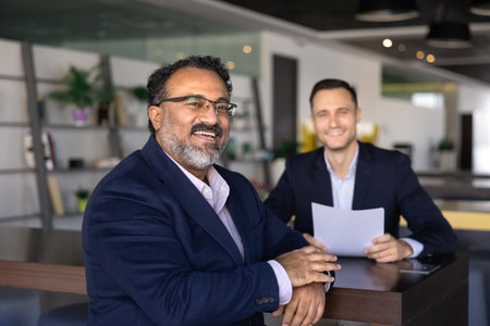 Happy senior Indian candidate posing for portrait after successful interview, sitting at table with young employer, human resource professional, getting job, smiling, looking at cameraの写真素材