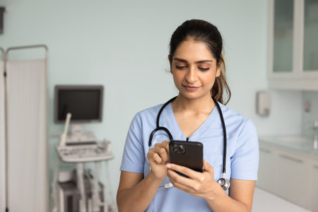 Focused young Indian nurse woman typing on mobile phone in hospital exam room. Female doctor in...