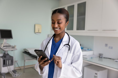 Smiling African female nurse in uniform using medical apps on smartphone standing in modern...