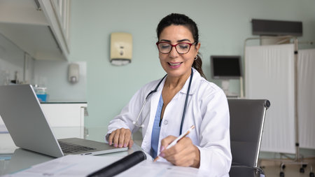 Middle-aged therapist woman in white coat working at desk in clinic office, making notes in journal, writing prescription or medical report, doing paperwork, perform administrative or clinical tasksの写真素材