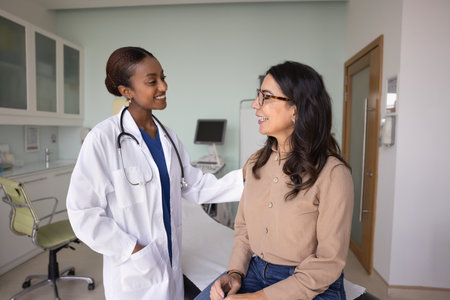 Support, positive communication. African female doctor and elderly 60s pensioner clinic client enjoy conversation in clinical office, gp put her hand on patient shoulder as gesture of encouragementの写真素材
