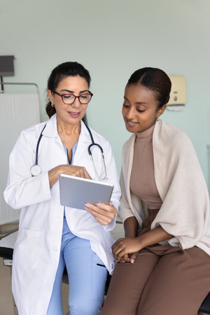 Elderly 60s female physician in white coat holds digital tablet, consulting young African woman patient during clinical visit. Doctor presents medical information, showing new tele-health applicationの写真素材