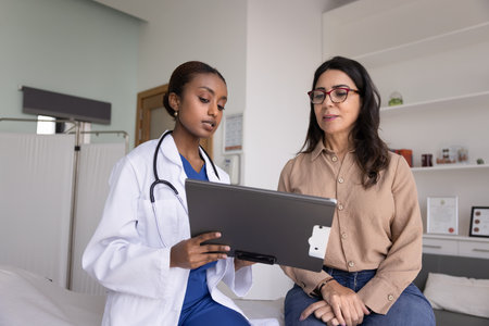Young African woman GP doctor in coat presents information on clipboard to older female patient during professional clinical consultation. Medical explanation, diagnostics review, treatment planningの写真素材