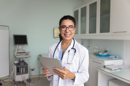 Smiling older 60s female physician standing in modern examination room with tablet, prescribing...